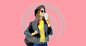 Stylish modern young woman drinks coffee wearing black rock leather jacket, sunglasses, round hat on pink studio background