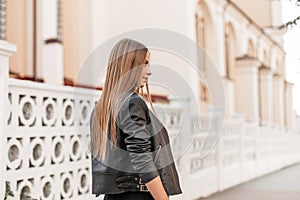 Stylish modern young woman with a long brown hair in trendy black leather jacket posing on a street in the city of near vintage