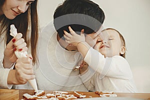 Stylish mother, father and cute little daughter decorating christmas gingerbread cookies with icing in modern room. Happy family