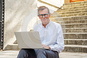 Stylish old man working on laptop surfing the internet sitting on stairs outdoors city in digital nomad Senior using modern