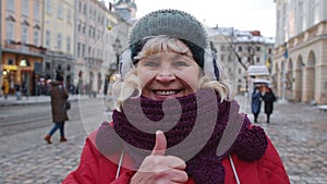 Stylish old senior woman tourist smiling, showing thumb up in winter city center of Lviv, Ukraine