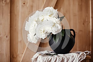 Stylish peony bouquet in black clay vase on linen fabric with scissors on rustic wooden background. White peonies rural still life
