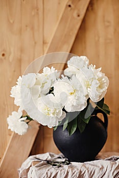 Stylish peony bouquet in black clay vase on linen fabric with scissors on rustic wooden background. White peonies rural still life