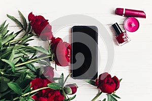 stylish phone with empty screen and beautiful red peonies, lipstick,nail polish and candle on white wooden background flat lay. s