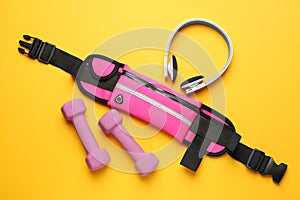 Stylish pink waist bag, dumbbells and headphones on yellow background, flat lay
