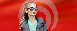 Stylish portrait beautiful blonde woman wearing a black rock jacket and sunglasses on red background