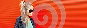 Stylish portrait of beautiful blonde woman wearing a black rock jacket and sunglasses on red background