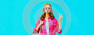 Stylish portrait of beautiful young woman blowing her red lips sending sweet air kiss wearing pink jacket, sunglasses on blue