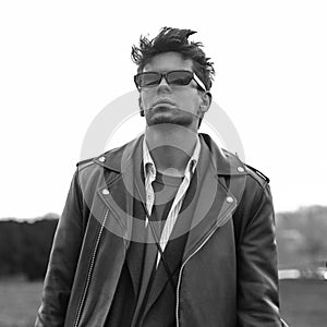 Stylish portrait cool young hipster man in fashionable dark sunglasses in vintage leather black jacket with trendy hairstyle