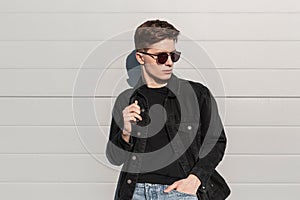 Stylish portrait urban young man with hairstyle in fashionable black jeans wear in trendy sunglasses with backpack near wall on