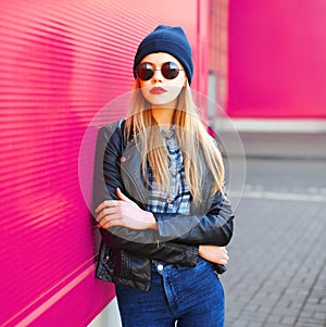 Stylish portrait young blonde woman in rock black style jacket, hat posing on city street over colorful pink wall