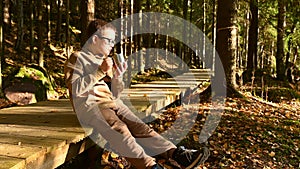 Stylish portrait of young boy sitting in the sunlight in autumn forest drinking tea.