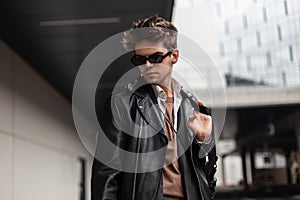 Stylish portrait of a young man hipster in sunglasses with a fashionable hairstyle in trendy black clothes near gray building on