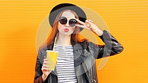 Stylish portrait of young woman with cup of juice wearing a black round hat, sunglasses over orange background