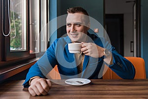 A stylish, positive businessman of Caucasian appearance, a man in a jacket and shirt sits at a table in a cafe on a coffee break