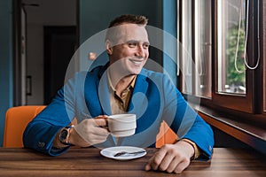 A stylish, positive businessman of Caucasian appearance, a man in a jacket and shirt sits at a table in a cafe on a coffee break