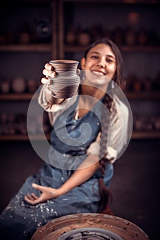 Stylish potter master woman siting on bench with pottery wheel and making clay pot. Folk handicraft.