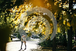 Stylish pregnant couple holding hands on baby bump and kissing under autumn leaves in sunny light. Happy young parents, mom and