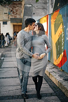 Stylish pregnant couple holding hands on baby bump and walking in city street. Happy young parents, mom and dad, hugging belly and