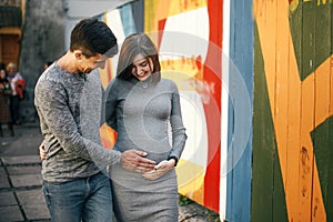 Stylish pregnant couple holding hands on baby bump and walking in city street. Happy young parents, mom and dad, hugging belly and