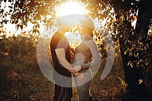 Stylish pregnant couple holding hands on belly and embracing in sunny light in autumn park under tree. Happy young parents, mom