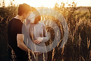 Stylish pregnant couple holding hands on belly and kissing in sunny light in autumn park. Happy young parents, mom and dad,