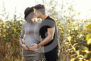 Stylish pregnant couple holding hands on belly in sunny light in autumn park. Happy young parents, mom and dad, hugging baby bump