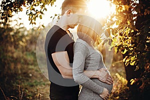 Stylish pregnant couple holding hands on belly in sunny light in autumn park. Happy young parents, mom and dad, hugging baby bump