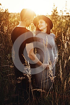 Stylish pregnant couple holding hands on belly in sunny light in autumn park. Happy young parents, mom and dad, hugging baby bump