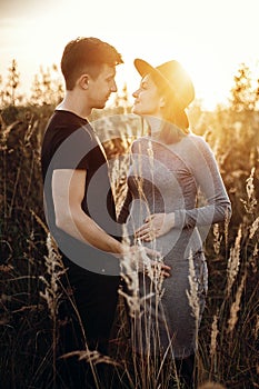 Stylish pregnant couple holding hands on belly in sunny light in autumn park. Happy young parents, mom and dad, hugging baby bump