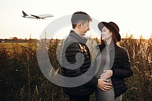 Stylish pregnant couple holding hands on belly in sunny light in autumn park. Happy young parents, mom and dad, hugging baby bump