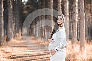 Stylish pregnant woman 25-30 year old wear knitted sweater holding her belly in woods outdoor.