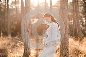 Stylish pregnant woman 25-30 year old wear knitted sweater holding her belly in woods outdoor.