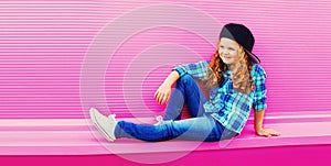 Stylish preteen curly girl sitting in baseball cap on pink background