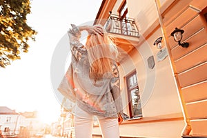 Stylish pretty modern blonde girl in fashion jeans outfit standing on the street and looking at sunset