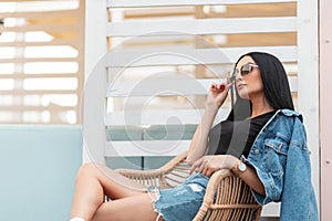 Stylish pretty young hipster woman in sunglasses in a black top in a vintage denim jacket is sitting on a straw chair in a cafe