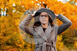 Stylish pretty young woman fashion model straightens chic hat. Attractive pretty trendy hipster girl in elegant coat with glasses