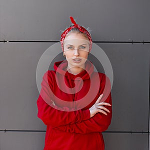 Stylish pretty young woman in modern fashionable clothes with a red bandanna is standing near a gray modern building