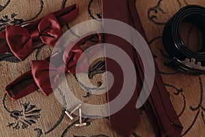 Stylish red bow-tie and tie with belt and cufflinks on bed. morn