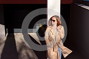 Stylish red hair woman standing at the urban streets in long beige coat and sunglasses