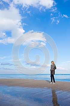 Stylish red-haired girl in dark coat and sunglasses standing on seaside with beautiful sky and water on background