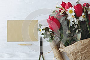 Stylish red tulips, daffodils bouquet with greeting card, pencil, scissors, twine on rustic wood