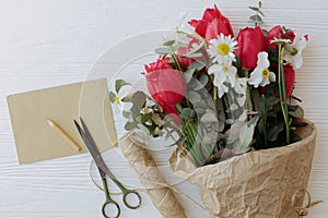 Stylish red tulips, daffodils bouquet with greeting card, pencil, scissors, twine on rustic wood