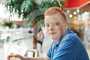 A stylish redhead hipster with freckles dressed in casual blue shirt standing in shopping mall near big green palm tree looking po