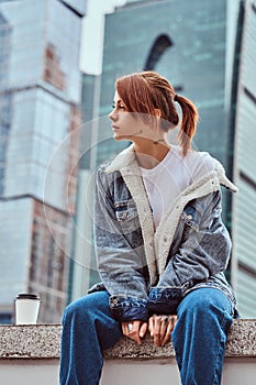 Stylish redhead hipster girl with tattoo on her face wearing denim jacket sitting in front of skyscrapers in Moskow city