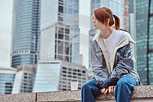 Stylish redhead hipster girl with tattoo on her face wearing denim jacket sitting in front of skyscrapers in Moskow city