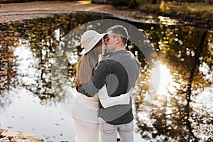 Stylish romantic young loving couple embracing and kissing by the pond at autumn park. Happy woman in hat and man