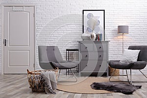 Stylish room interior with grey chest of drawers and chairs near white brick wall