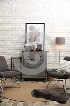 Stylish room interior with grey chest of drawers and chairs near white brick wall