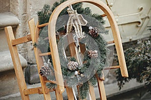 Stylish rustic wreath on wooden sleigh at building exterior. Modern christmas decor in city street. Winter holidays in Europe.